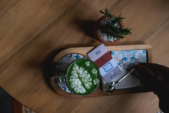 Overhead view of a teak table: a matcha latte with leaf latte art on a patterned tile coaster, a wooden board with a Malu card and a Breathe card, a hand reaching for a spoon, and a small potted plant