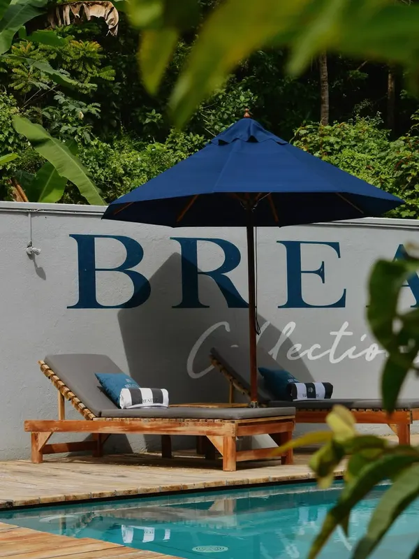 Two teak sun loungers with Breathe cushions under a navy parasol beside the pool, against the wall painted with the Breathe Collections name