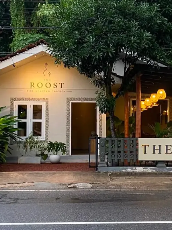 The Roost seen from across the road on an overcast day: the rooster mark and name painted on the gable, two windows framed with patterned tiles, and amber lanterns lit on the veranda
